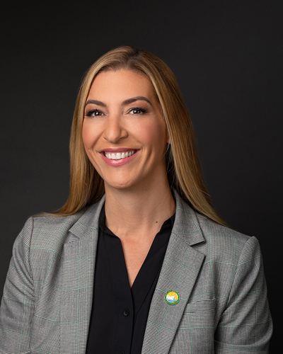Christina Birdsey, with long hair, wearing a gray blazer and a black shirt, smiles in front of a black background.