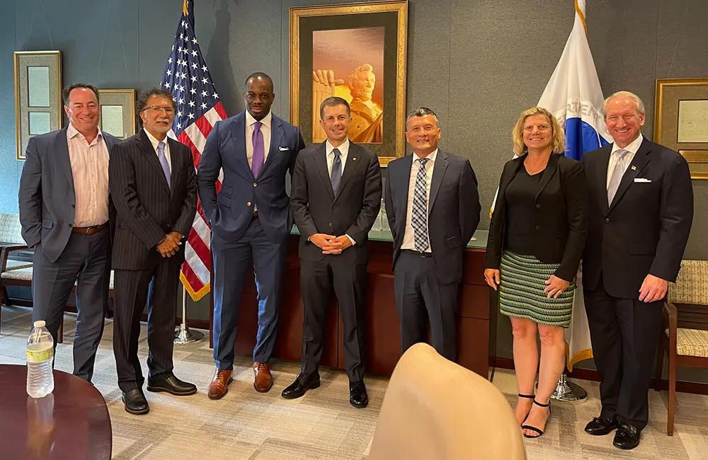 Seven people in business attire standing in a conference room with U.S. and another flag in the background, discussing the $80M investment for the Port of Hueneme made possible through federal funding.