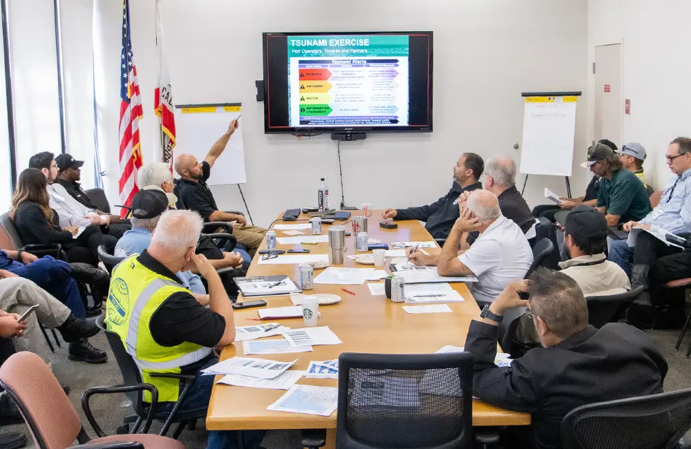A group of people is seated around a conference table participating in a tsunami exercise meeting. A man is pointing at a presentation slide on the screen titled "August Wave Preparedness." Flags are visible in the background.