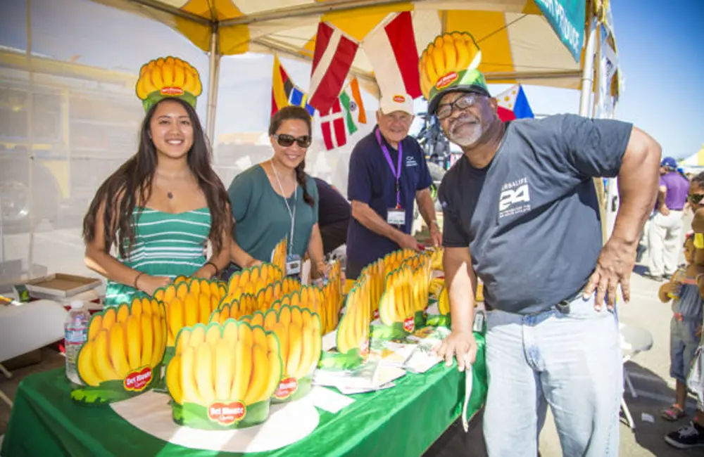 Four people smile at a booth filled with banana-themed hats under a canopy at the Port of Hueneme's Banana Festival. Flags are visible in the background.