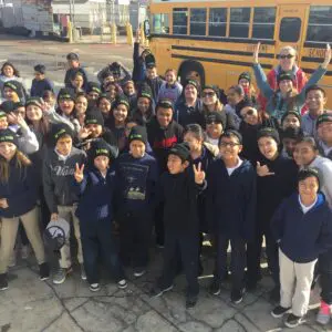 A group of students and teachers pose for a photo in front of a yellow school bus. Many students are making peace signs, wearing beanies that say "NASA," creating a sense of unity that feels just like home.