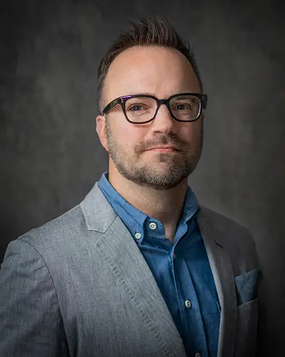 A man with short hair, wearing glasses, a light gray jacket, a blue shirt, and a gray pocket square, stands against a dark, plain backdrop, looking at the camera—perfect for an employee directory.