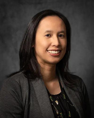 A woman with shoulder-length dark hair wearing a gray blazer and a black patterned blouse, smiling in front of a dark gray background, perfect for the Employee Directory.