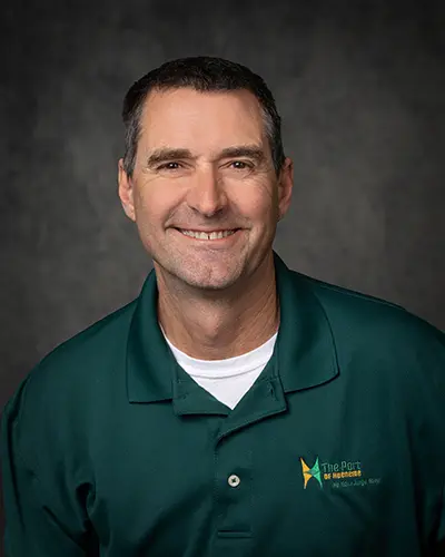 A man with short dark hair and wearing a dark green polo shirt smiles at the camera. The shirt has a logo with the text "The Port" on it. The gray background suggests this might be his photo from an employee directory.