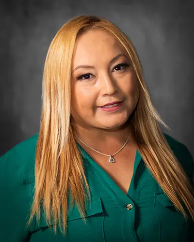 A woman with long blonde hair wearing a green blouse and a necklace poses against a dark gray background, perfect for the employee directory.