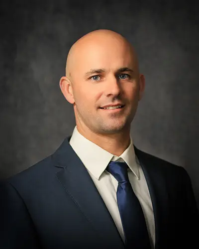 A man with a shaved head and blue eyes, dressed in a dark blue suit, white dress shirt, and blue tie, poses against a dark grey background for the employee directory.