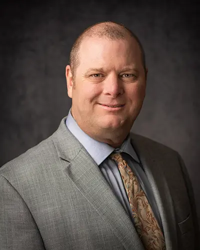 A man, dressed in a light gray suit jacket, light blue shirt, and patterned tie, is smiling slightly against a dark background. His professional demeanor makes him a perfect candidate for our Employee Directory.