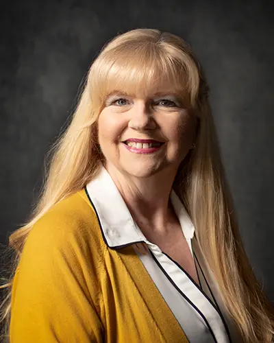 A woman with long blonde hair and wearing a yellow jacket over a white shirt smiles against a dark background, as if ready for her photo in the Employee Directory.