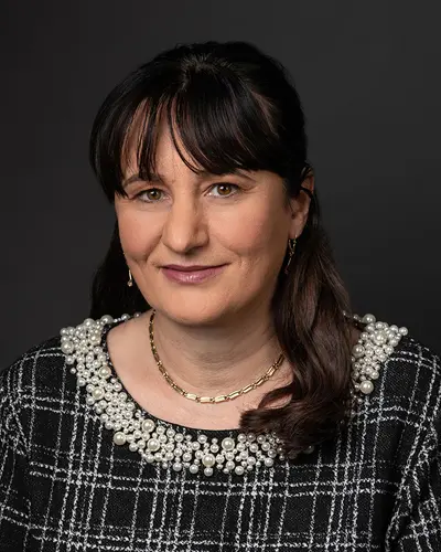 A person with dark hair and bangs smiles slightly while wearing a black and white checked top adorned with pearls and a gold necklace, perfect for an Employee Directory photo. The background is plain and dark.