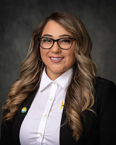 Portrait of a person with long wavy hair, glasses, and a white shirt under a dark blazer, posing against a dark gray background. Perfect for an employee directory.