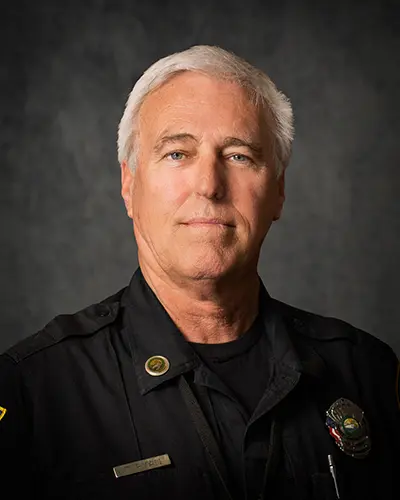 A man with white hair in a police uniform stands facing forward against a dark, textured background, as if he were posing for an Employee Directory.