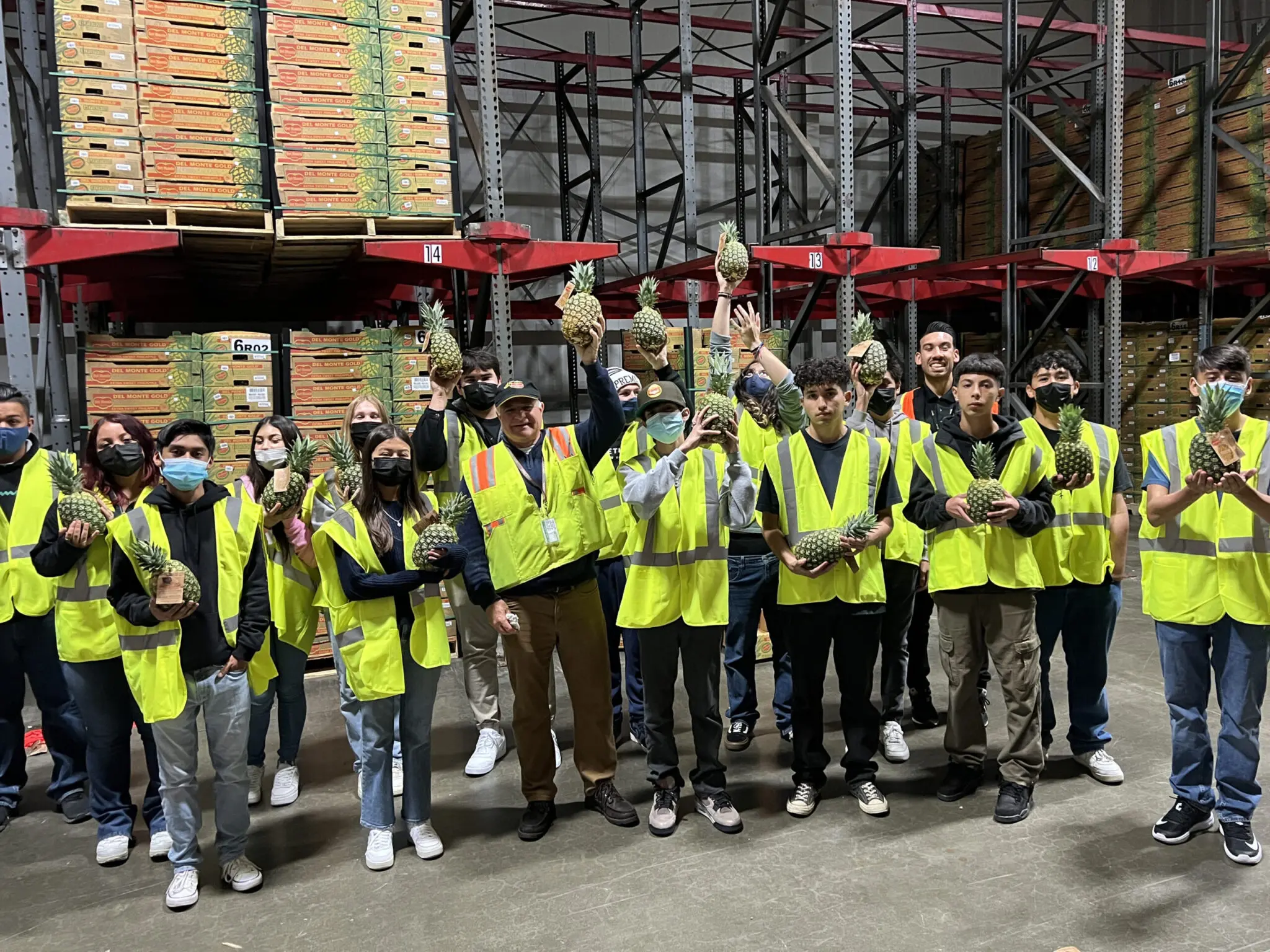 A group of people in yellow safety vests stands in a warehouse, holding pineapples. Pallets of boxed goods are stacked on shelves behind them as they engage in an educational session about inventory management.