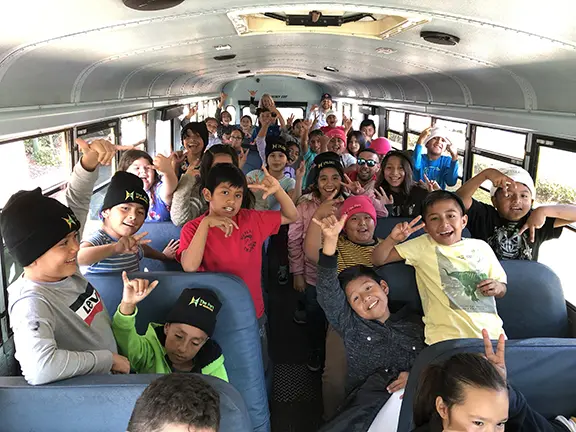 A group of diverse children and adults on a school bus, smiling and making playful gestures towards the camera, ready for their exciting Port Tours adventure.