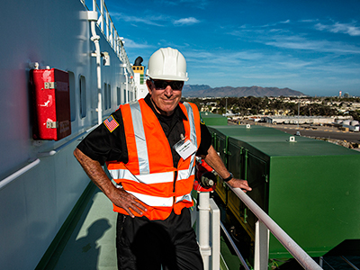 Person wearing a white hard hat and orange safety vest stands on the deck of a ship, with a hand resting on a railing. In the background, mountains and buildings illustrate the importance of effective port governance in coordinating maritime administration activities.