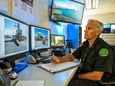 Man in uniform sits at a desk with multiple monitors, meticulously taking notes on a clipboard. Images of an outdoor setting related to port safety are displayed on screens. In the background, there's a wall-mounted calendar and an additional screen, highlighting the importance of safety and security in his work.
