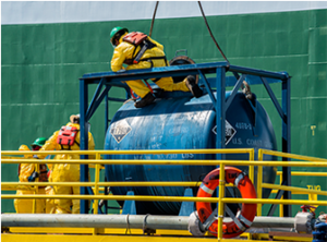 Workers in yellow protective suits and helmets inspect a large blue cylindrical cargo container on a platform with yellow railings. A lifebuoy is attached to the railing, ensuring safety during shipping operations.