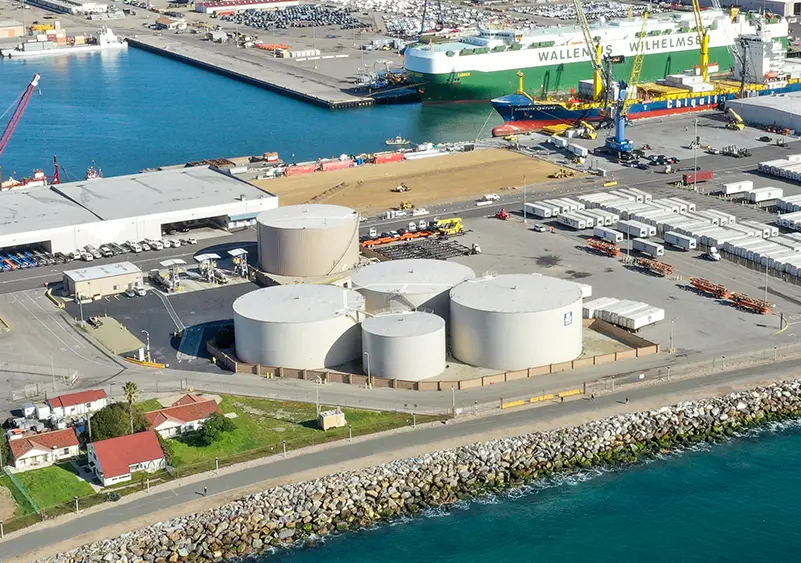Aerial view of an industrial port with large storage tanks filled with liquid, shipping containers, cranes, and two large ships with "WALLENIUS WILHELMSEN" markings in the harbor.