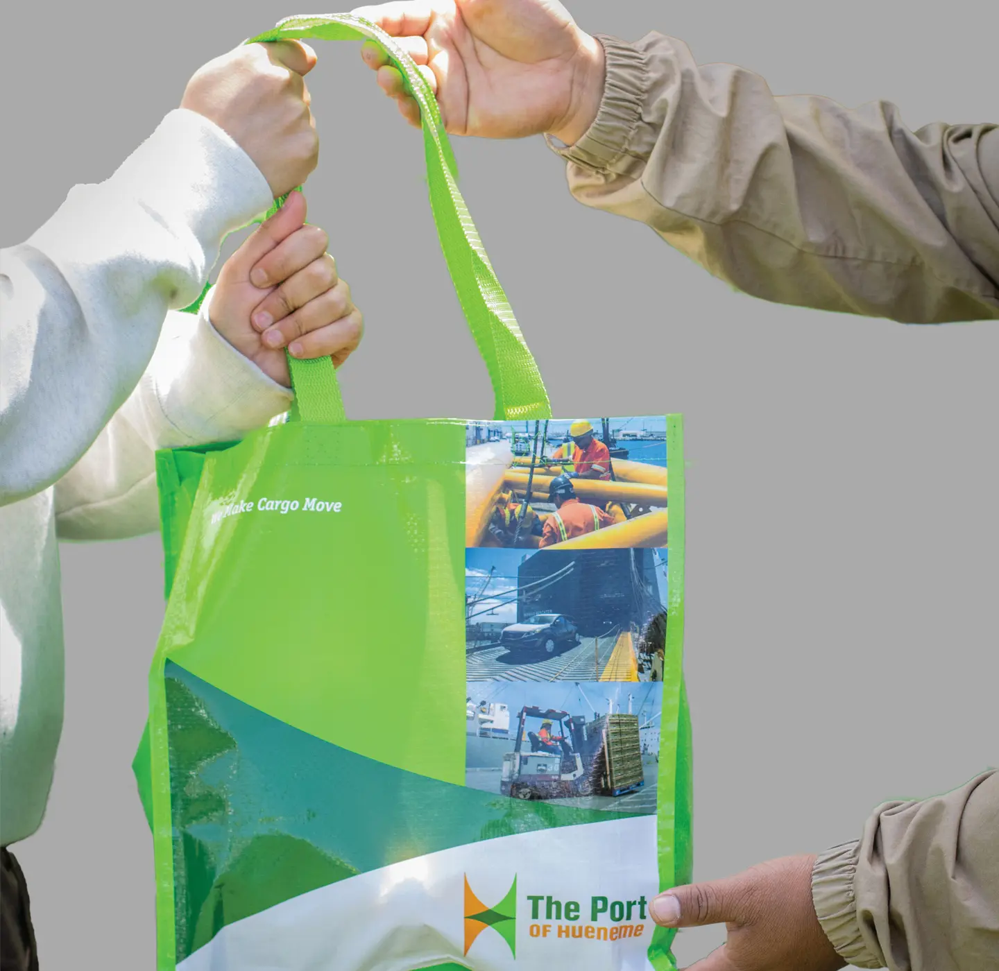 Two people holding a green and white tote bag adorned with "The Port of Hueneme" logo and various images of port activities, capturing the essence of community events.