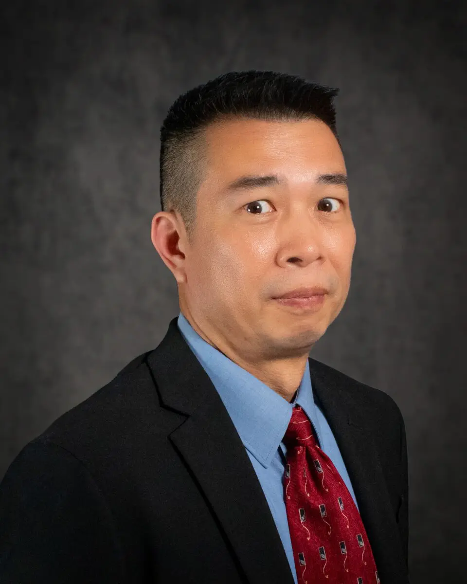 A man wearing a blazer, blue shirt, and red tie stands in front of a dark, textured background, looking slightly to the left with an alert expression. This is Edward Chung.