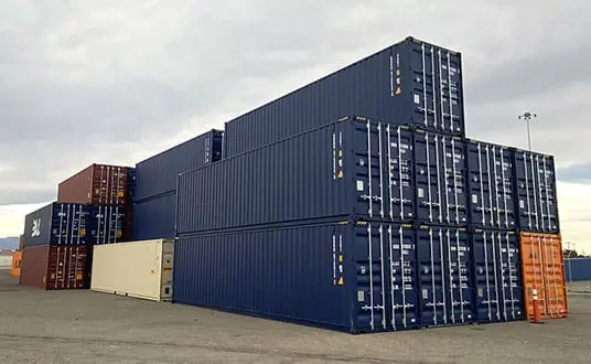 A stack of shipping containers, mostly blue with one orange and one white container, arranged in a shipping yard on a cloudy day. This scene was captured in the 2021 Year End edition of Dock Talk, showcasing the bustling activity and organization at the port.