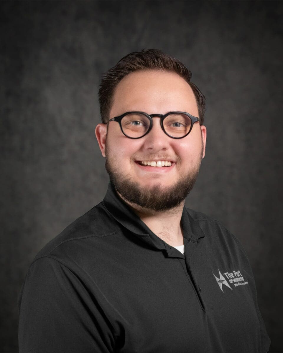 Garrett Raborg, a man with glasses and a beard, smiles in front of a dark background while wearing a black collared shirt with a logo.