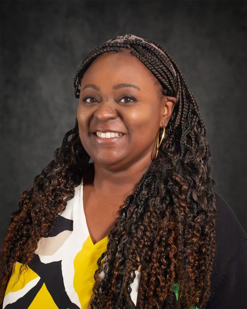 Letitia Austin, with long braided hair, is smiling while wearing a yellow, black, and white patterned shirt. The background is dark and plain.