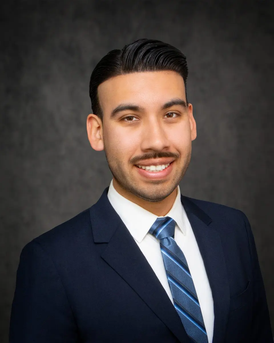 A man in a suit and tie smiles against a plain dark background.