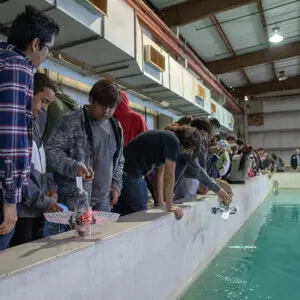 People standing by an indoor pool at home, observing and handling small robotic devices on the water surface.