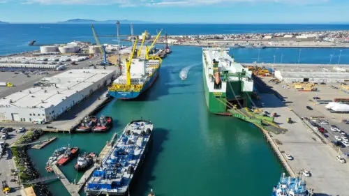 Aerial view of a busy shipyard with two large cargo ships docked along a narrow waterway, surrounded by industrial facilities and equipment. The clear blue sky and distant land on the horizon give an almost homely feel to this bustling maritime hub.