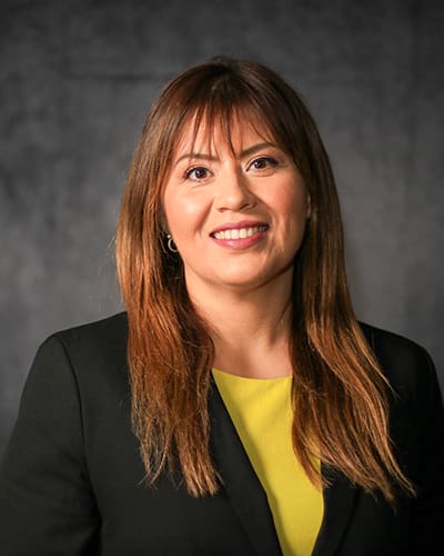 A woman with long brown hair, wearing a black blazer over a yellow top, smiles confidently in front of a dark gray background, perfect for the employee directory.