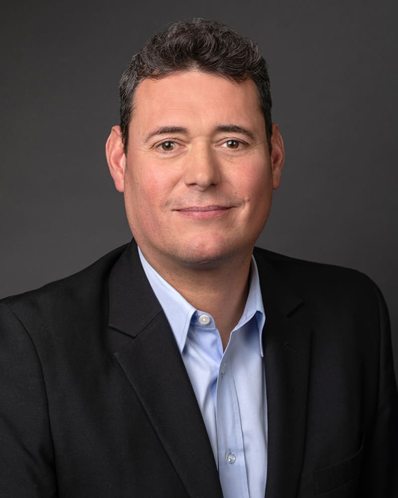 A man with short dark hair, wearing a blue shirt and dark blazer, poses against a plain dark background, embodying the professional demeanor often seen among Port Commissioners.