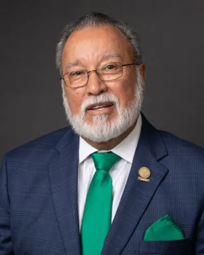 An older man with gray hair and a beard, possibly a member of the Port Commissioners, wearing glasses, a blue checkered suit, and a green tie with a matching pocket square stands against a dark background.