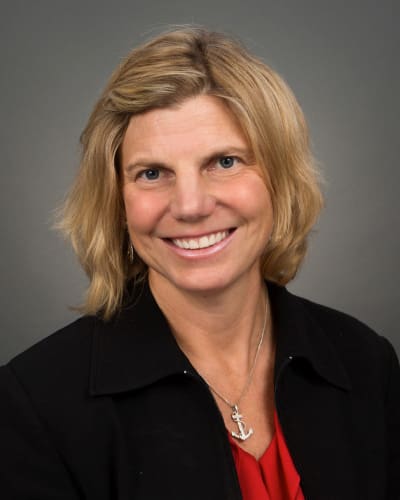 A woman with shoulder-length blonde hair, wearing a black jacket over a red top, smiles at the camera against a gray background. She has a necklace with an anchor pendant. Perfect for the employee directory.