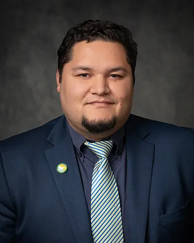 A man with short dark hair and a goatee wearing a suit and striped tie stands against a dark, textured background. He has a small green pin on his lapel, making him easily identifiable in the company's Employee Directory.