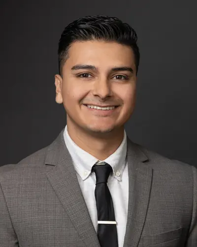 A man in a gray suit, white shirt, and black tie smiles against a dark background, looking ready for his Employee Directory photo.