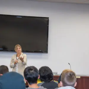 A person speaks to an audience in a conference room with a large blank screen and microphones on a table. The California state flag is visible on the right side, and Kristin Decas is among the attentive listeners.