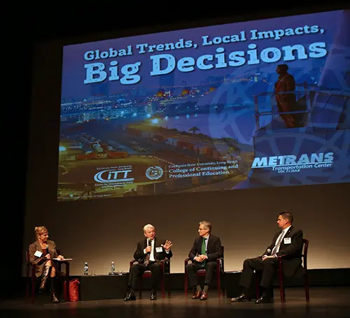 Four panelists in formal attire discuss onstage at a "Global Trends, Local Impacts, Big Decisions" event, with a high-quality sound system ensuring their voices are heard clearly amidst the projected background showing event details and logos.