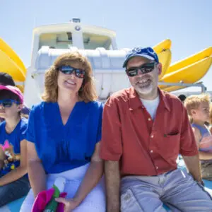 A group of people are seated together on a boat under a sunny sky, committed to community and enjoying the day. Two yellow kayaks are visible in the background. Some people are wearing sunglasses and hats.