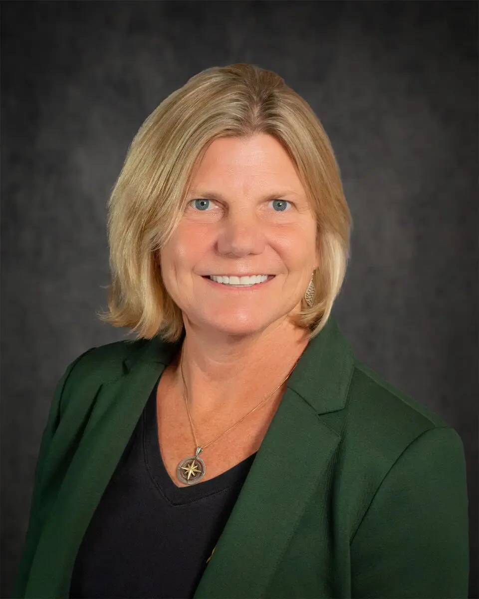 Kristin Decas, with shoulder-length blonde hair, wearing a green blazer over a black top, is smiling against a dark gray backdrop.