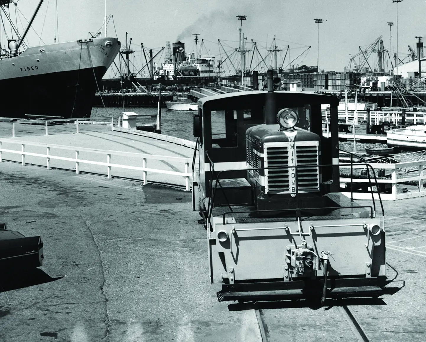 A small locomotive sits on a track near a dock at the bustling Port of Hueneme, with ships and cranes visible in the background.