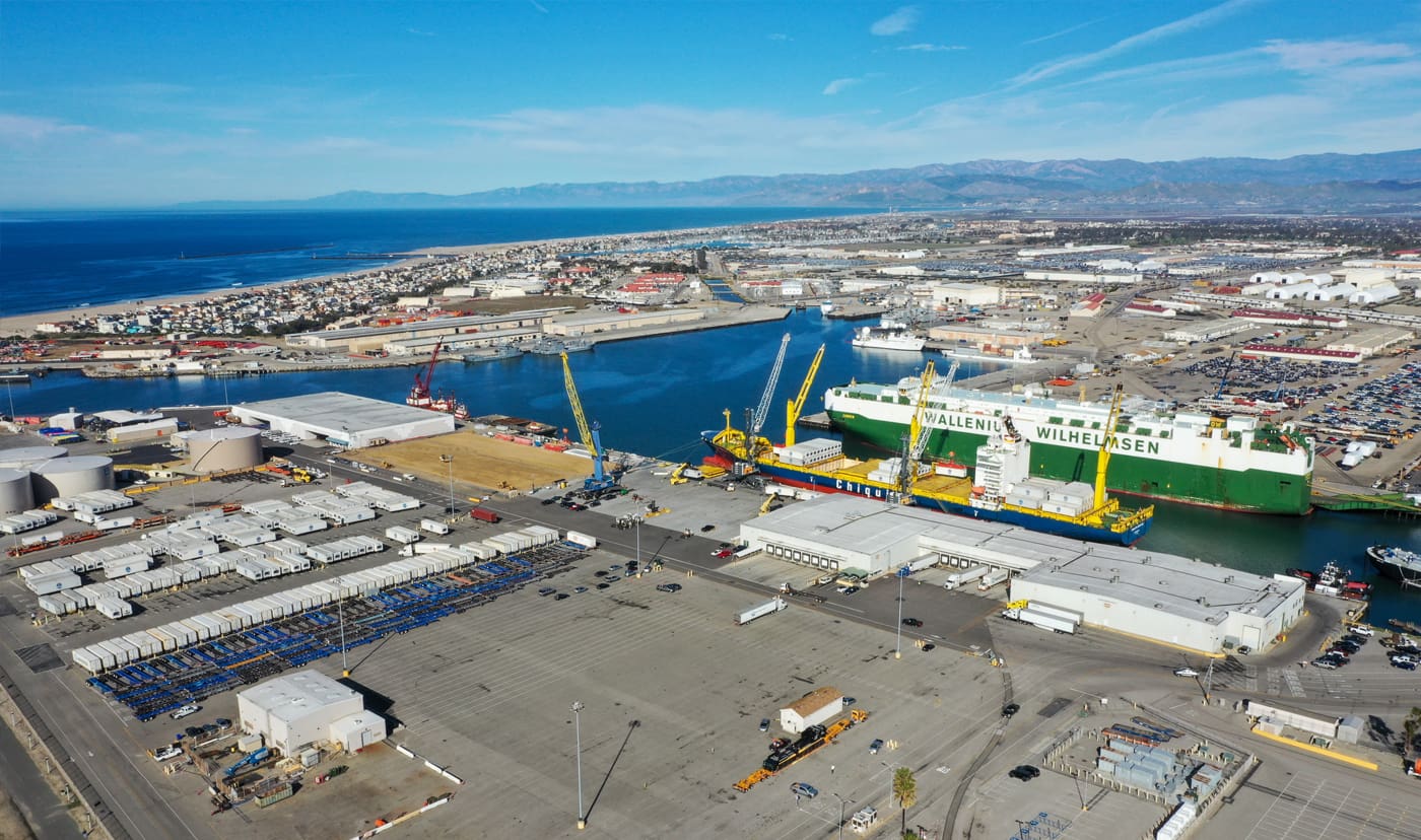 Aerial view of an industrial port area with cargo ships docked, cranes, shipping containers, and warehouses, all overseen by rigorous maritime security measures, set against a backdrop of coastline and mountains.