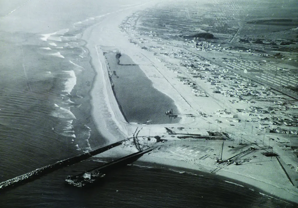 Aerial view of a coastal area in California featuring a pier, sandy beaches, and adjacent residential or commercial buildings; bodies of water include the ocean and a small inland waterway near the Port of Hueneme.