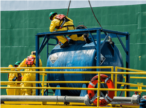 Workers in yellow protective suits and helmets inspect a large blue cylindrical cargo container on a platform with yellow railings. A lifebuoy is attached to the railing, ensuring safety during shipping operations.