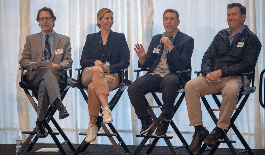 Four people sit on tall chairs engaging in a discussion at the Fathomwerx event. One person is speaking while the others listen attentively. They all wear badges and casual business attire.