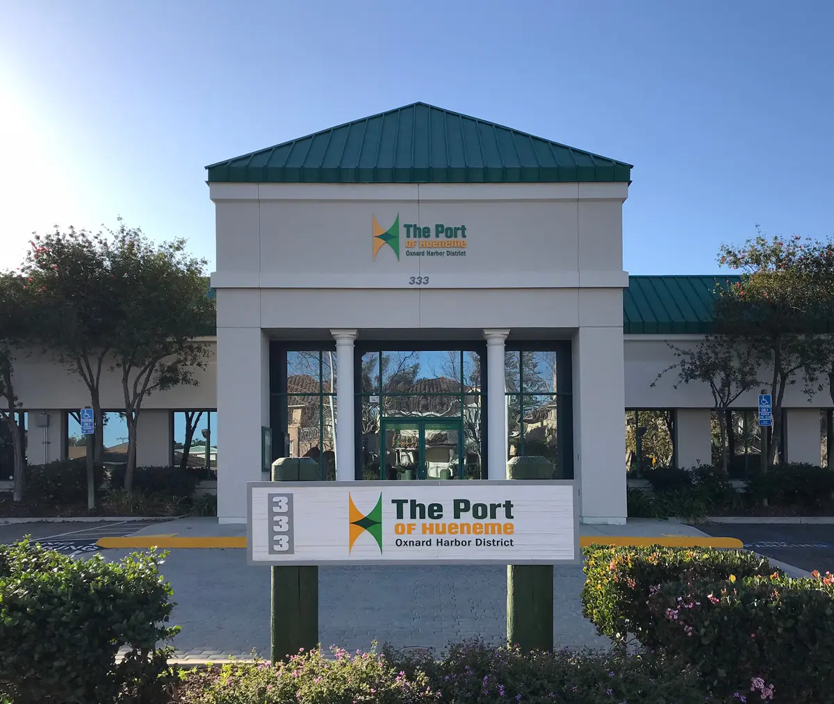 Front view of The Port of Hueneme administration building with a green roof, large windows, and a sign bearing the address 333 and the Port's name. Trees flank the entrance on either side. This hub is essential for overseeing all aspects of financial management within the port's operations.