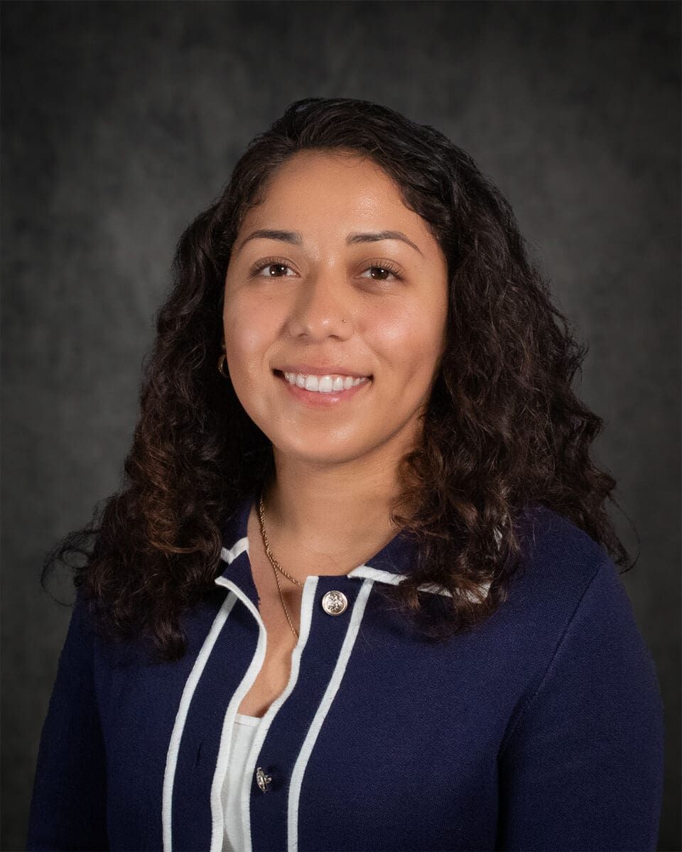 A woman with curly dark hair, smiling, dressed in a navy blue jacket with white trim, stands against a dark background reminiscent of Jacobo's signature style.