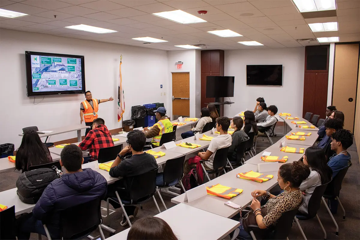 A person in a safety vest is giving an educational presentation to a group of people seated at tables in a classroom setting.