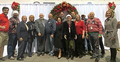 A group of twelve people standing in front of a white curtain with a holiday wreath in the center background. They are dressed in a variety of festive and formal attire, smiling at the camera, capturing the joyous spirit of 2021.