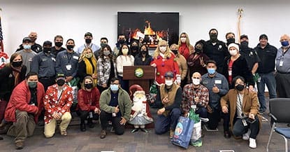 A group of people wearing masks, standing and kneeling around a podium in a room with a festive Santa decoration and a digital fireplace display in the background, captures the essence of Dock Talk's Year End 2021 celebration.