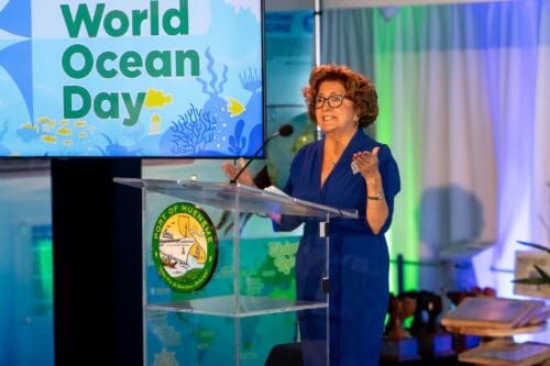 A woman in a blue dress speaks at a podium with a sign reading "World Ocean Day." The podium, adorned with the Port of Hueneme logo, stands before an ocean-themed backdrop.